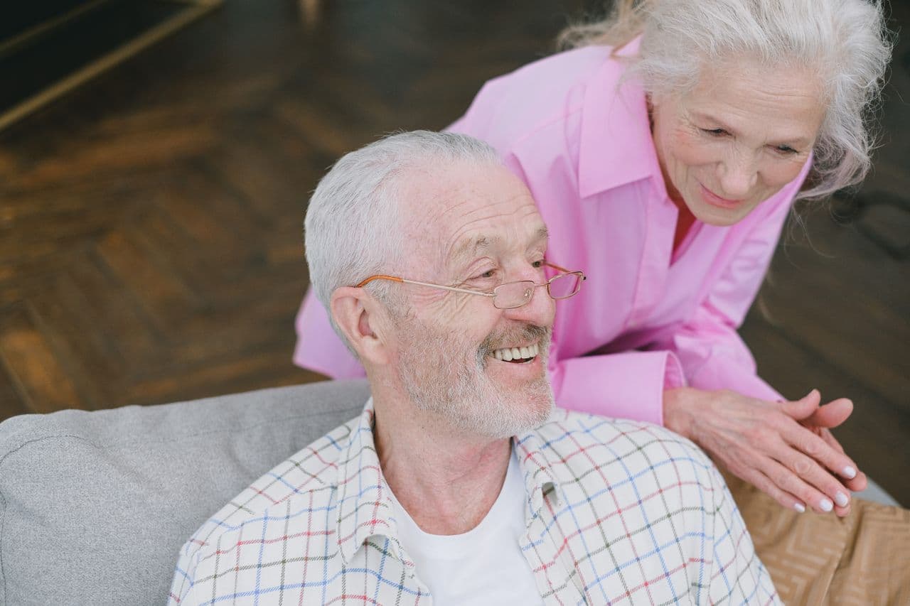 An older man smiling on a sofa while a woman in a pink shirt leans beside him